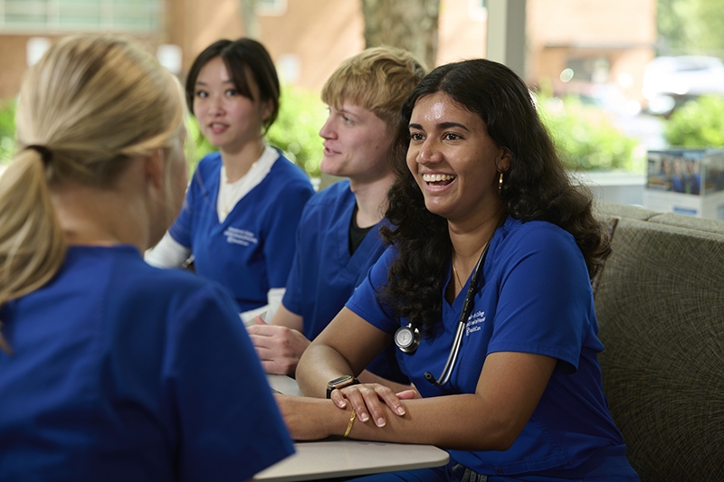 Student Sradha Ajithkumar in a blue scrub uniform sitting with other students smiling and laughing.