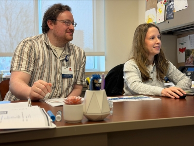 Brian Chervitz works with program director Kristen Neils sitting down in front of a desk.