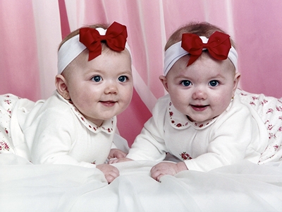 Twins Katie and Kelsey Boyer as babies with a red bow and white outfits.