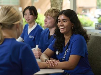 Student Sradha Ajithkumar in a blue scrub uniform sitting with other students smiling and laughing.